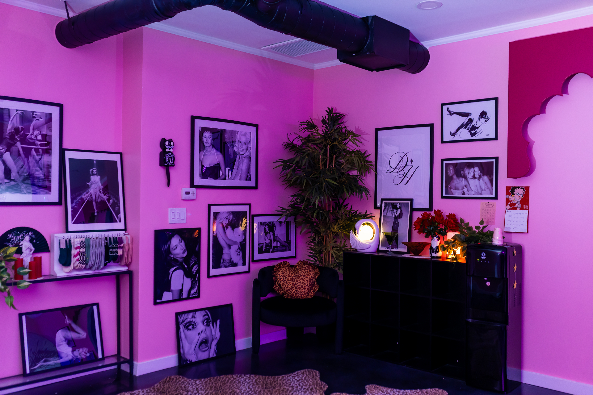 Pink studio wall covered in framed black-and-white pinup and fashion prints beside a black chair